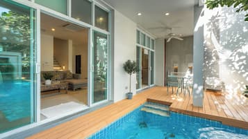 Una piscina interior moderna con agua azul cristalina, rodeada por una terraza de madera y puertas de cristal que dan a una sala de estar con un sofá y una mesa de comedor.