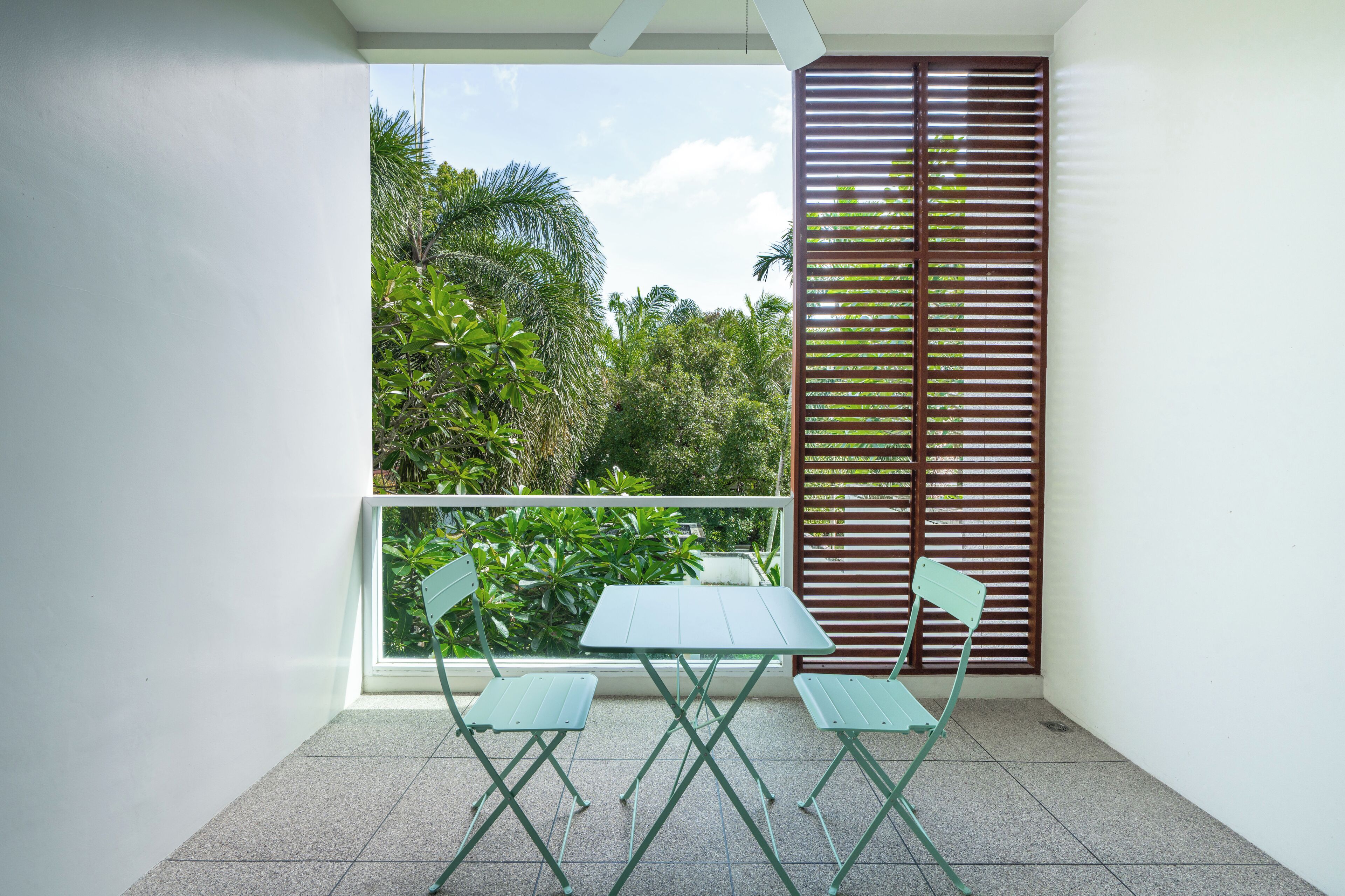 A balcony with a table and chairs, a view of greenery, and a window with blinds.