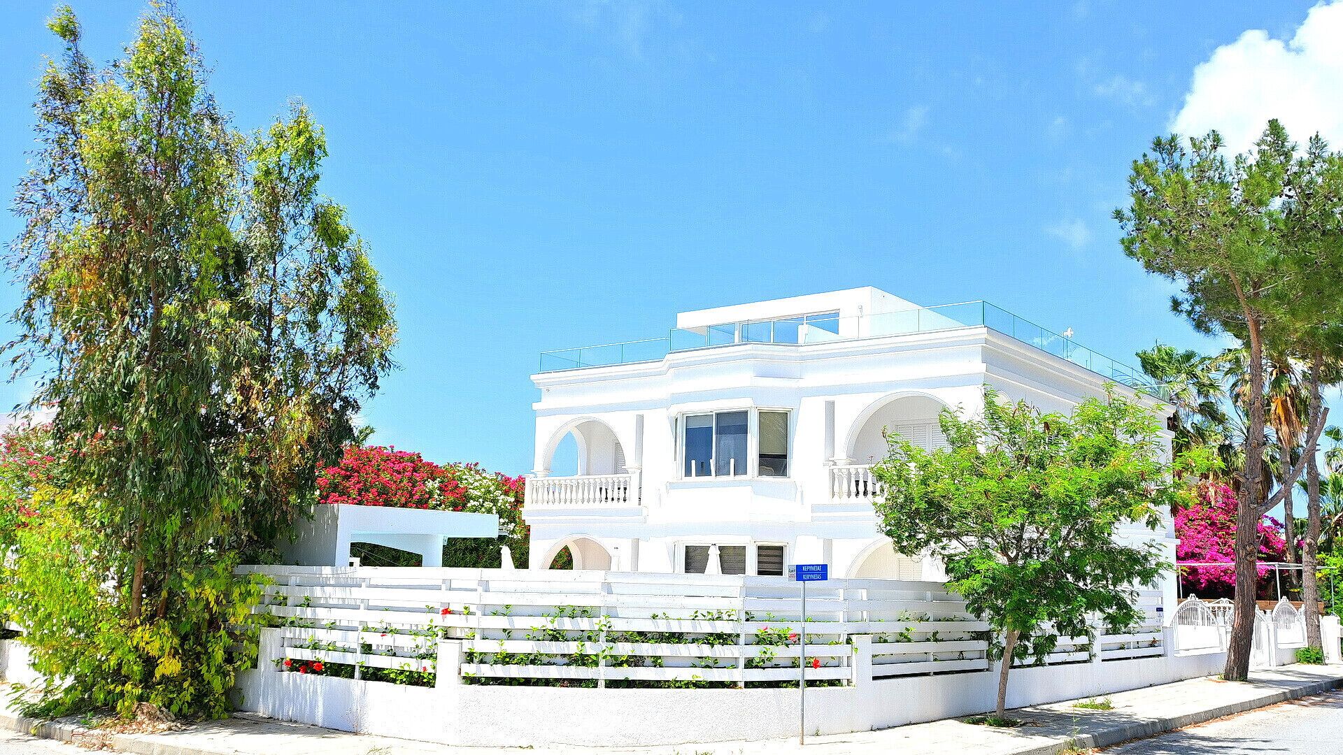 A white building with a balcony, surrounded by greenery and flowers.