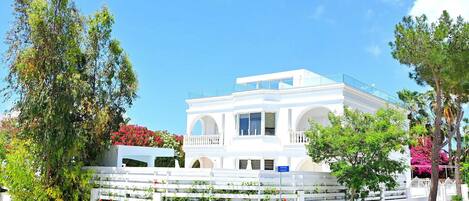A white building with a balcony, surrounded by greenery and flowers.