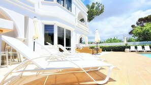 A poolside area with lounge chairs and umbrellas, a building with large windows, and a view of the surrounding greenery. - Villa Cecilia Coral - Beautiful Mansion in Coral Bay (Coral Bay)