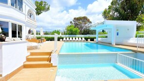 Une piscine dotée de chaises longues et d’un espace abrité, entourée d’un bâtiment blanc et de verdure.