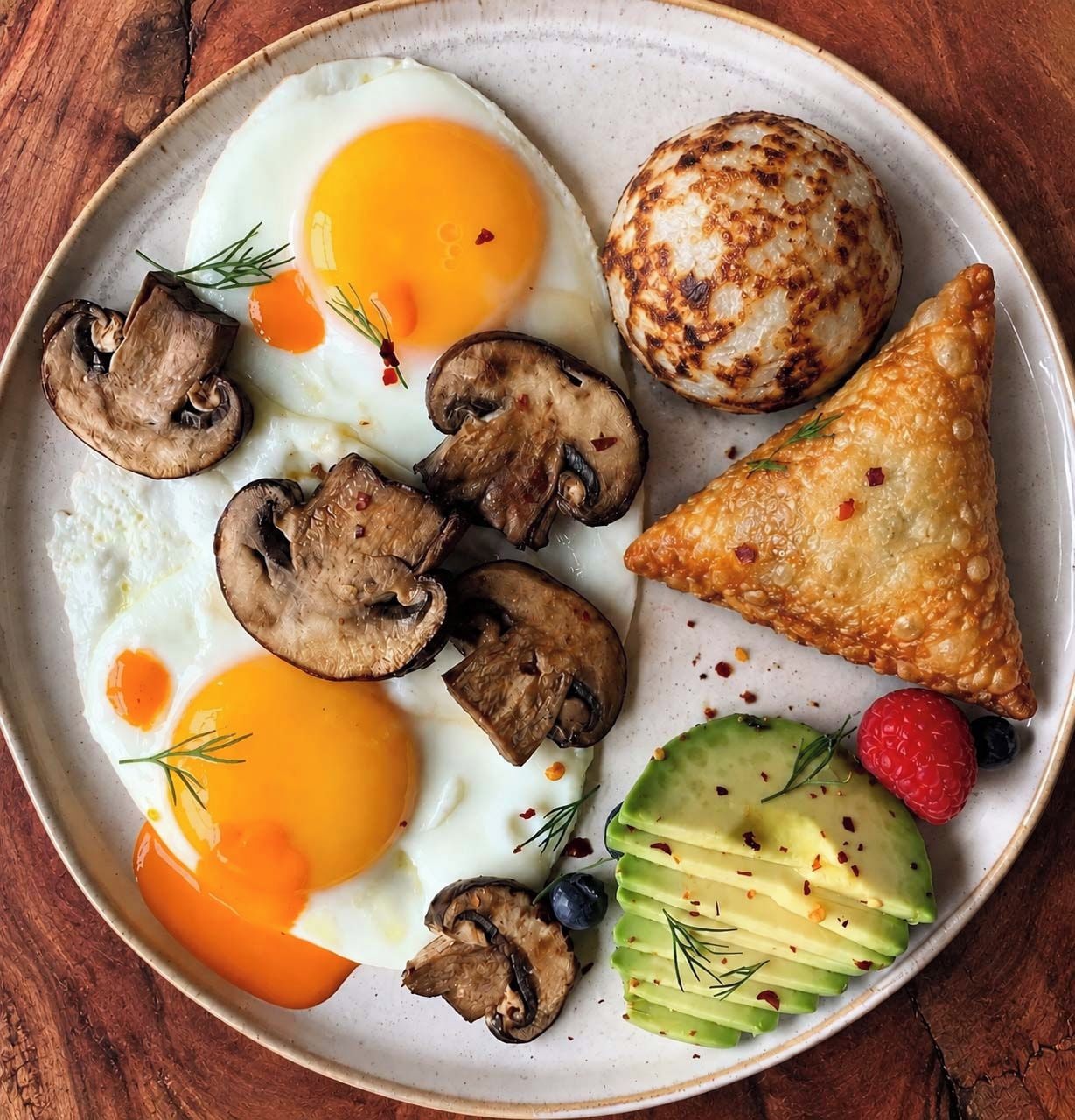 Un plato con dos huevos fritos, champiñones a la plancha, una patata al horno, un pastel triangular, aguacate en rodajas, una frambuesa y un arándano.