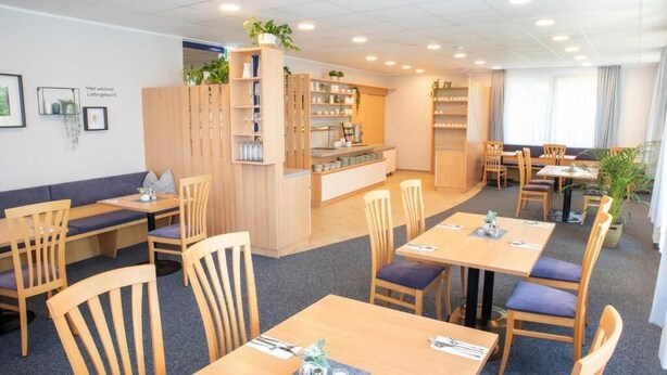 Comedor de hotel con mesas y sillas de madera, una recepción y estanterías con plantas.