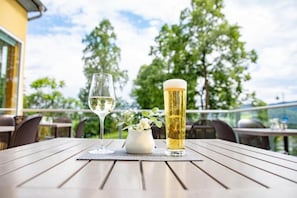 A table with a glass of white wine and a tall glass of beer, a small vase with flowers, and a view of trees and a building in the background.