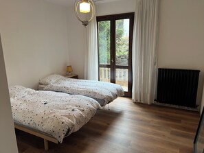 Ein Schlafzimmer mit zwei Betten, einem Nachttisch, einer Lampe, einem Fenster mit Vorhängen und einer Heizung.