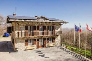 A stone building with a balcony, solar panels, and flags. - An entire house made of stone and wood, surrounded by greenery (Bagnolo Piemonte)