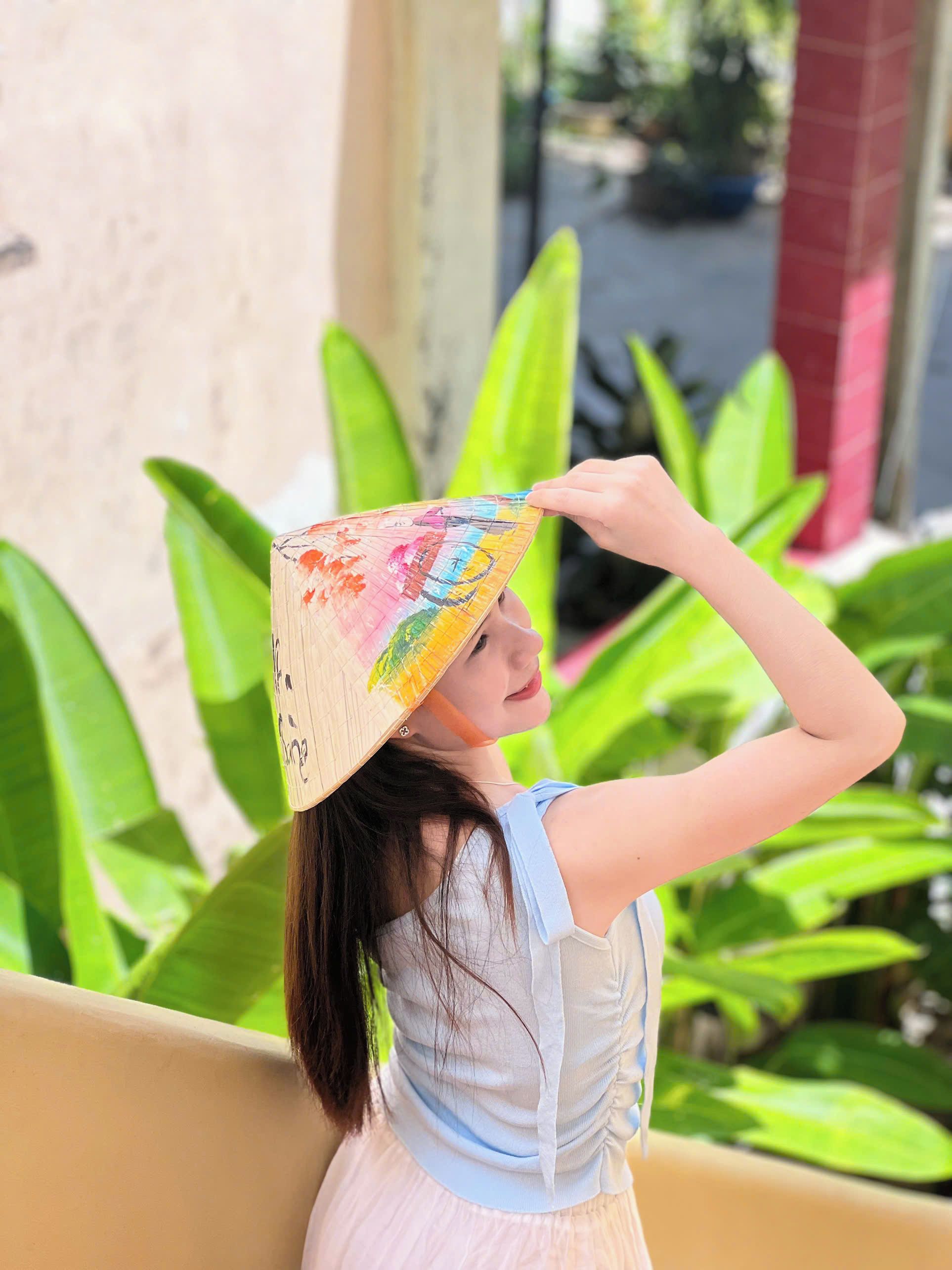 A person holding a colorful hat with a pattern, standing outdoors with green foliage in the background.