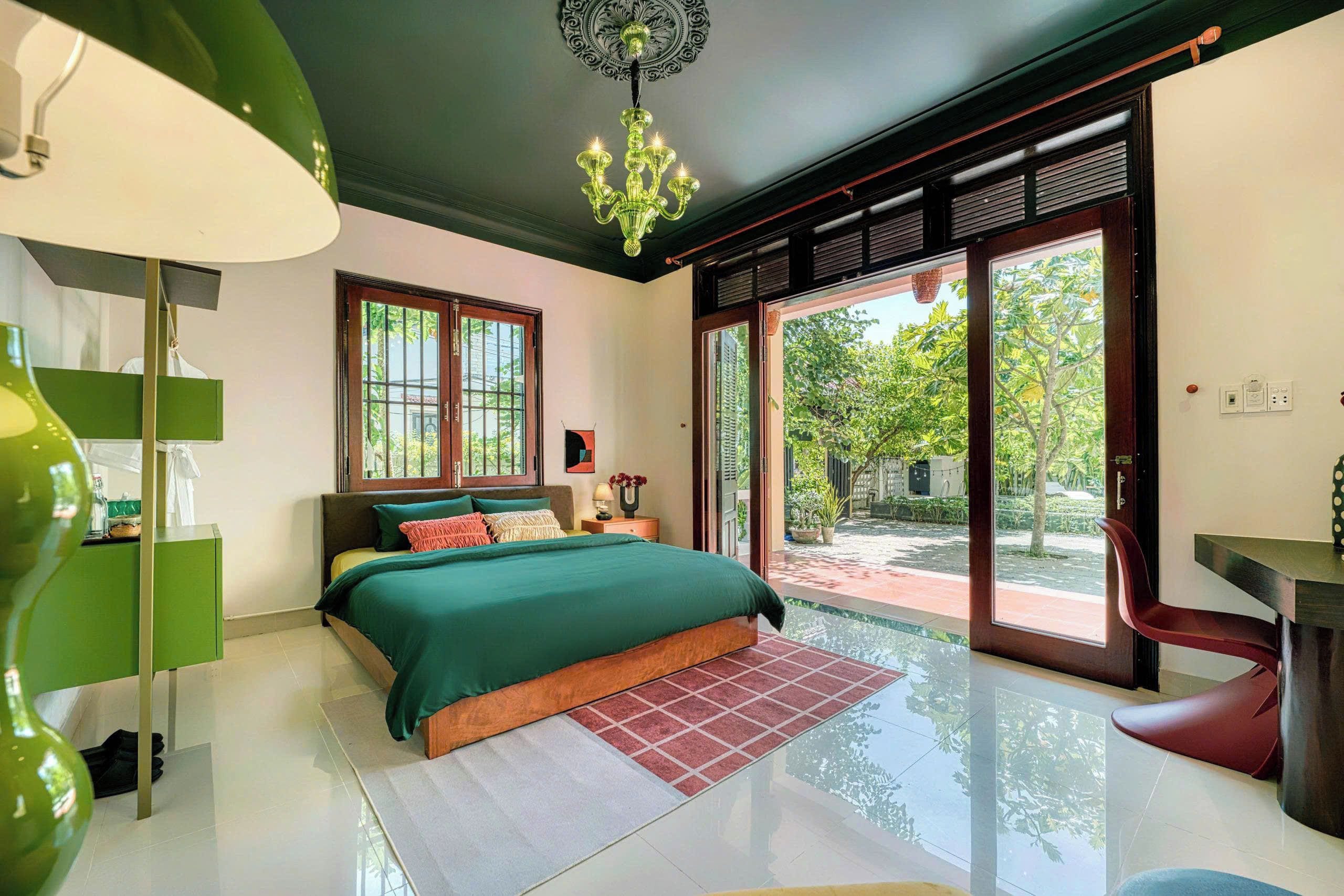 A bedroom with a bed, a desk, a chair, a chandelier, and a view of the outdoors.