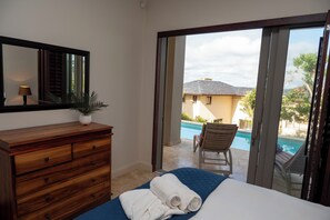 A bedroom with a wooden dresser, a mirror, a potted plant, a bed with a blue bedspread and white towels, and a view of a swimming pool and a building through a glass door. - Silver Horizon (Knysna)