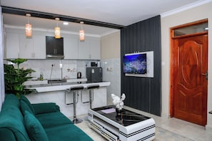 A modern kitchen with white cabinets, a black countertop, and a built-in microwave. There is a green sofa and a small table with a vase of flowers. - Stylish 1BR Apartment with Balcony Near JKIA & SGR (Nairobi)
