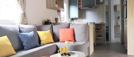 Ein modernes Wohnzimmer mit einem grauen Sofa, gelben und blauen Kissen, einem weißen Couchtisch mit Büchern und Tassen, und einer Küche im Hintergrund.
