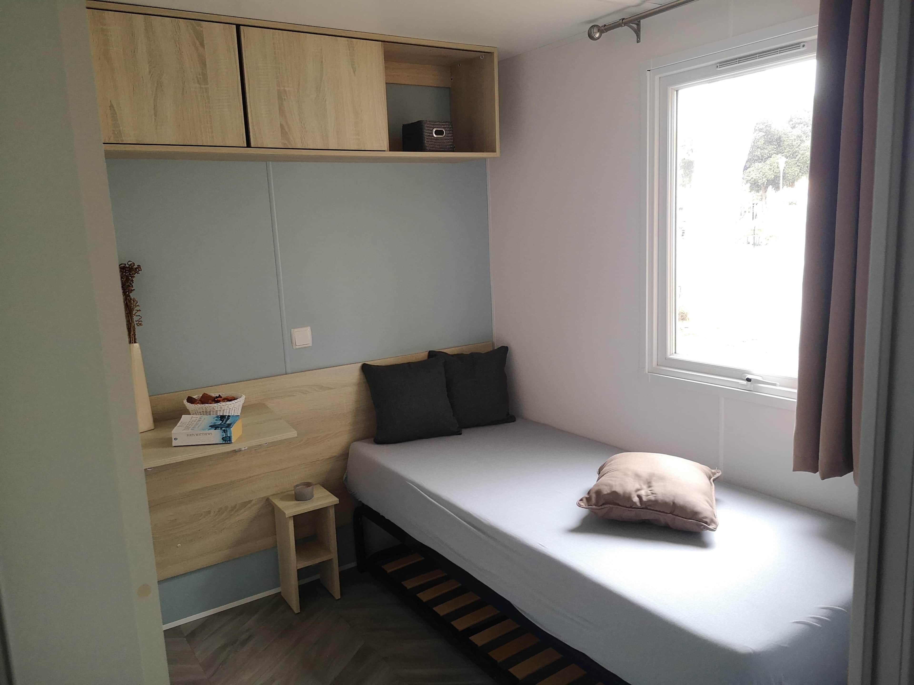 Une petite pièce, de taille modeste, comprenant un lit simple, une étagère en bois et une fenêtre.