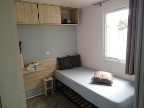 Ein kleines, schlichtes Zimmer mit einem Einzelbett, einem Holzregal und einem Fenster.