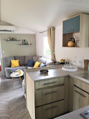 Un espacio habitable compacto con kitchenette, un sofá con cojines, una pequeña mesa de comedor y una ventana con cortinas.
