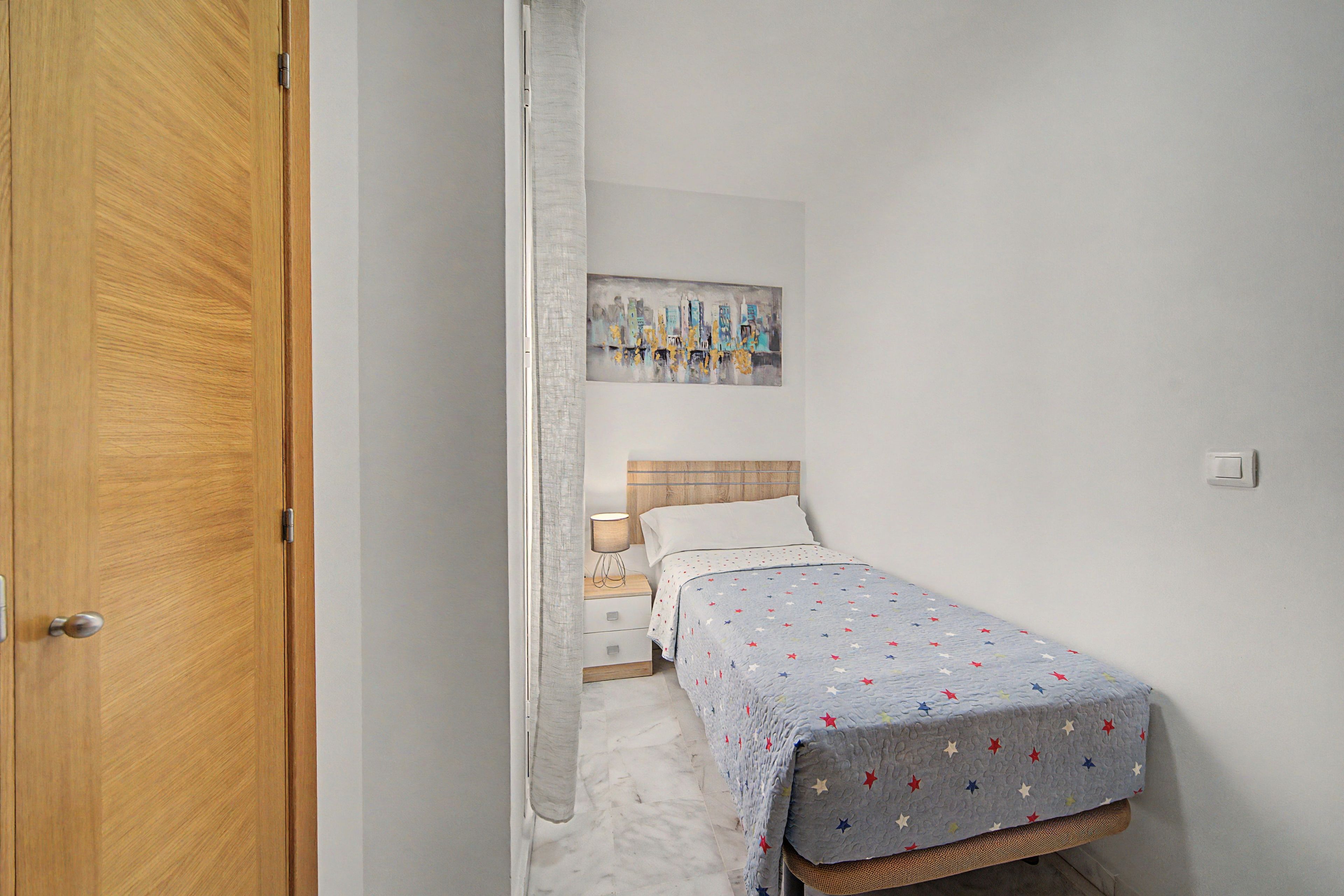 Une petite chambre à coucher, bien rangée, avec un lit simple, une table de chevet, un tableau accroché au mur et une porte.