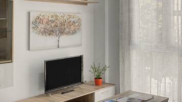 Un salon moderne avec un téléviseur à écran plat, un tableau représentant un arbre décoratif, une plante en pot et une table basse avec des magazines.