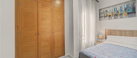 Un dormitorio con un armario de madera grande, una cama con colcha de estampado de estrellas, un cuadro en la pared y una mesita de noche con una lámpara.
