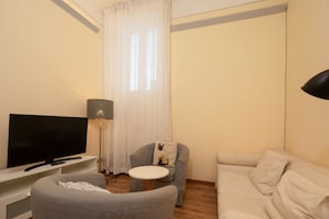 A living room with a flat-screen TV, a small round table, a grey armchair with a cushion, and a sofa. - Cavalleggeri Palace - 3-Bedroom Apartment (Firenze)
