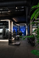 Un gimnasio con diverso equipo de ejercicio, una pantalla digital grande que muestra una imagen y un diseño arquitectónico moderno con conductos a la vista.