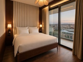 Habitación de hotel con una cama grande, dos lámparas de noche, vista a un paisaje urbano a través de un ventanal amplio y una lámpara de techo.