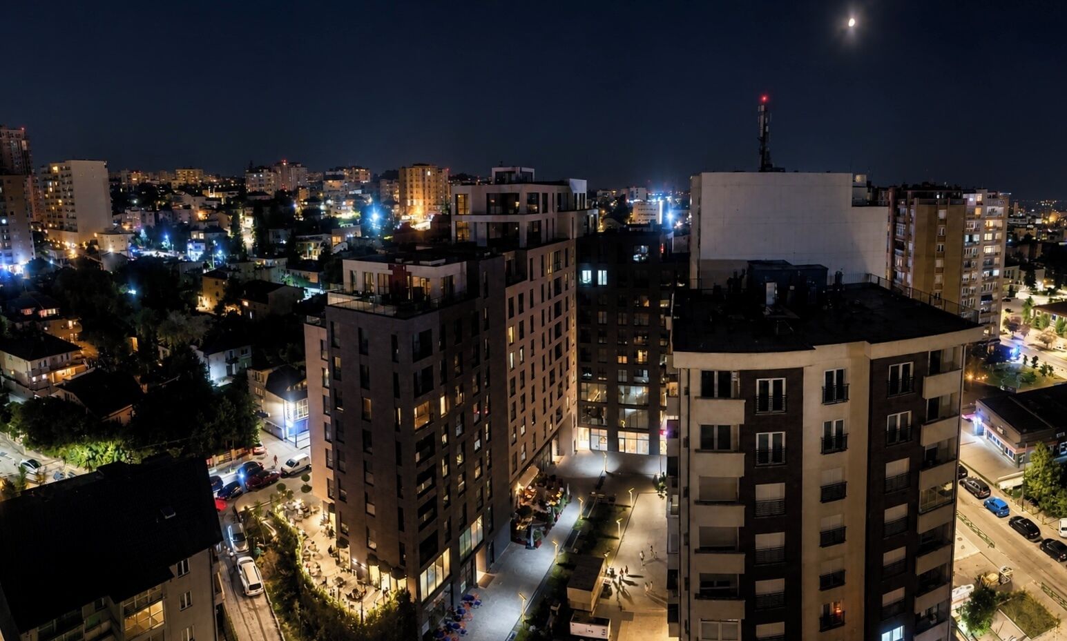 Paisagem urbana noturna com arranha-céus, janelas iluminadas e uma rua visível.
