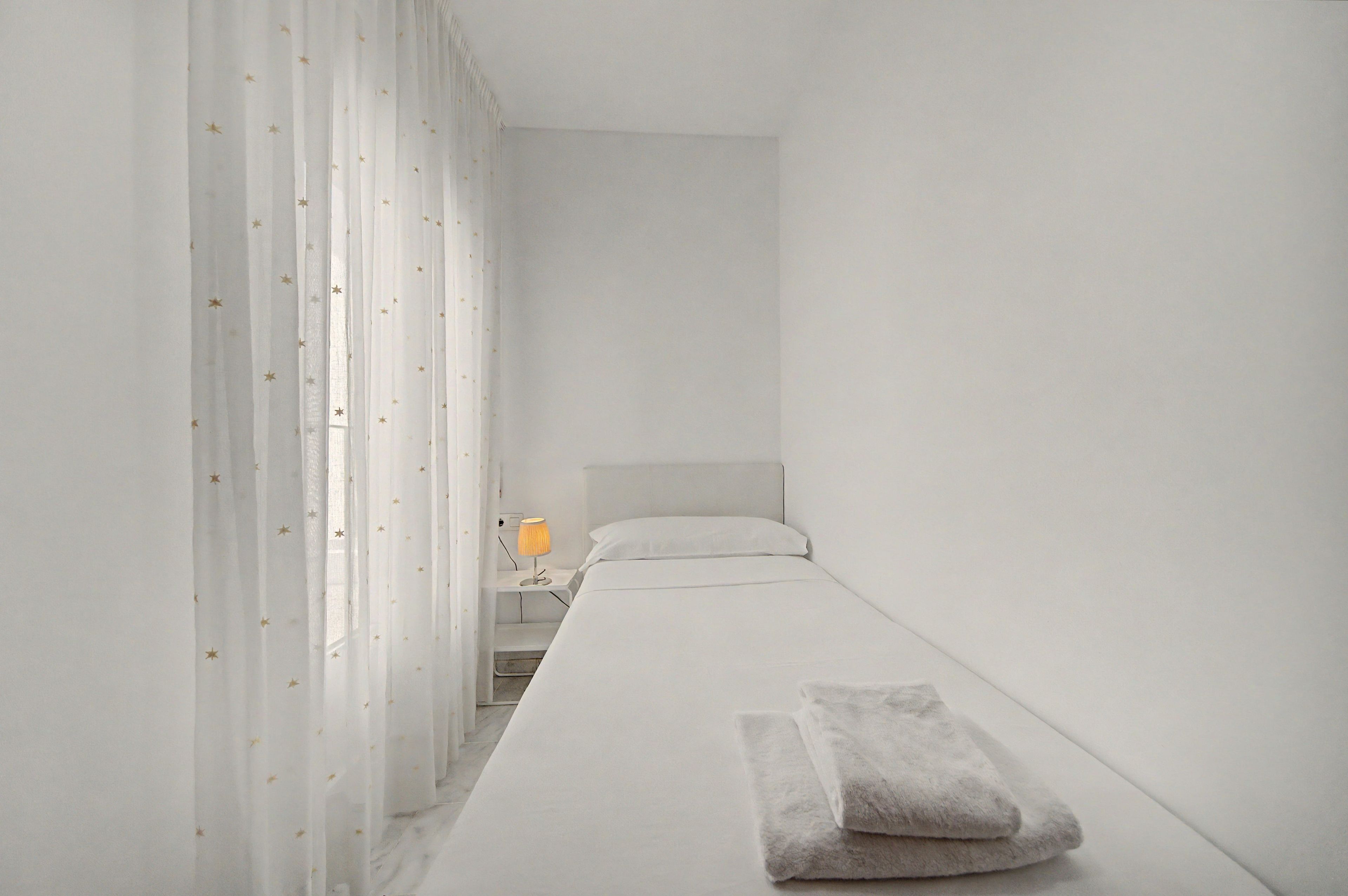 Une chambre minimaliste avec un lit simple, une table de chevet avec une lampe et des rideaux légers ornés de motifs d’étoiles.