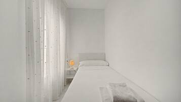 Une chambre minimaliste avec un lit simple, une table de chevet avec une lampe et des rideaux légers ornés de motifs d’étoiles.