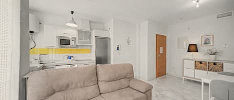 A modern living room with a beige sofa, a white dining table, and a kitchen area with a microwave and refrigerator.