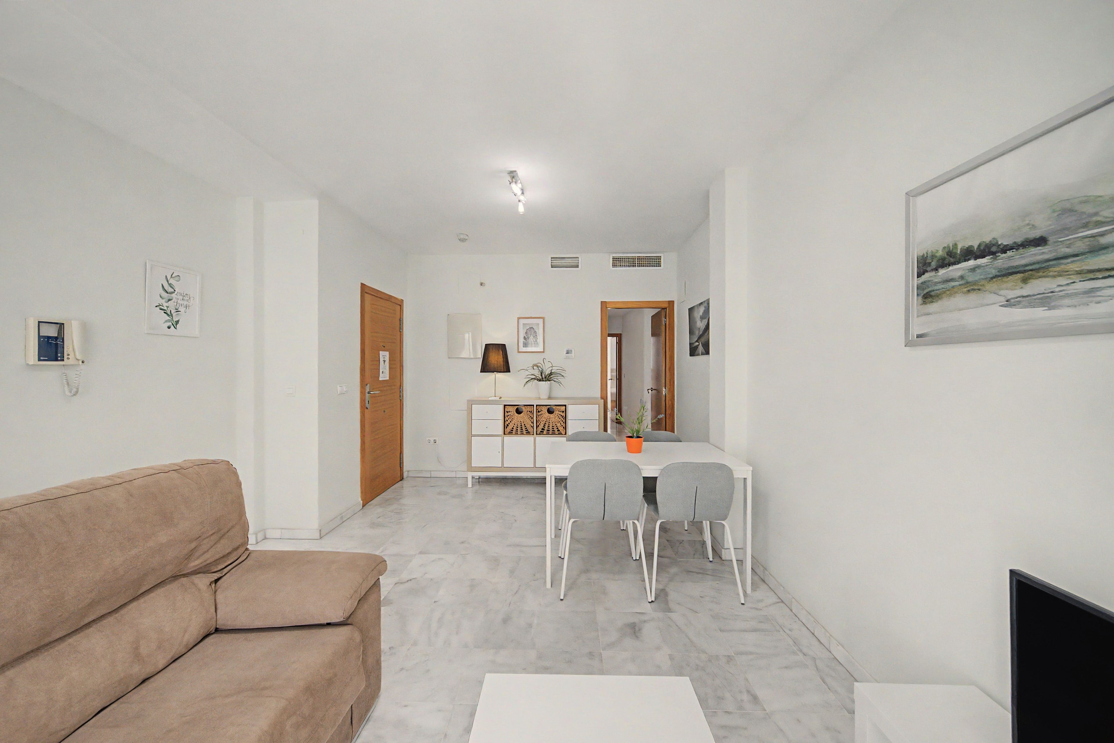 Un salon avec un canapé beige, un coin repas avec une table et des chaises blanches, et un tableau accroché au mur.