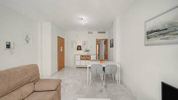 Un salon avec un canapé beige, un coin repas avec une table et des chaises blanches, et un tableau accroché au mur.