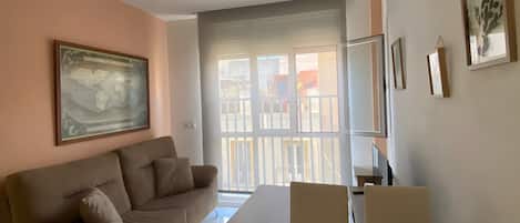 A living room with a sofa, a dining table, and chairs. There is a large window with a view of the outside.