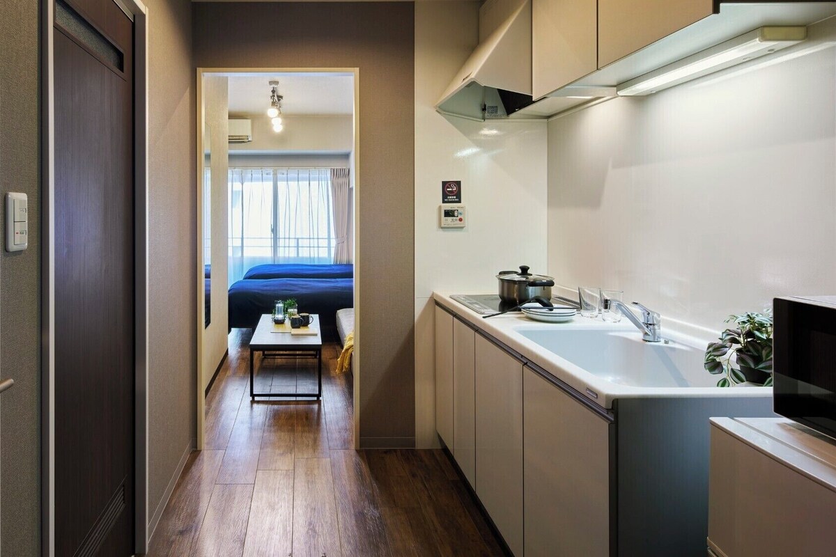 Un pasillo estrecho conduce a un dormitorio con una cama y una pequeña mesa con objetos encima. A la derecha se ve una kitchenette con un fregadero y una estufa.