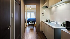 Un pasillo estrecho conduce a un dormitorio con una cama y una pequeña mesa con objetos encima. A la derecha se ve una kitchenette con un fregadero y una estufa.