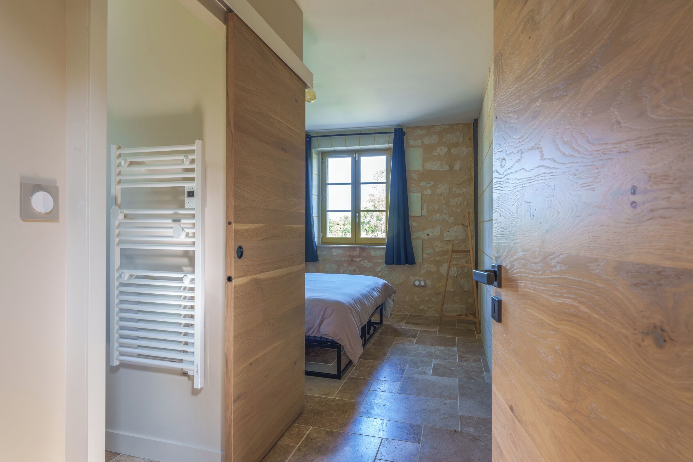 Un pasillo estrecho con un radiador, una cama con cabecera y una ventana con cortinas.