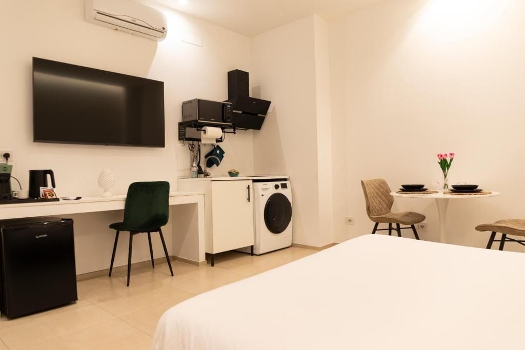 Kompakti hotellihuone, jossa on sänky, työpöytä tietokoneella, pesukone, pieni jääkaappi ja seinälle asennettu televisio.