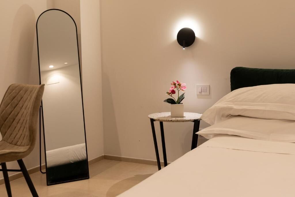 A bedroom with a bed, a chair, a round table with a plant, a wall-mounted light, and a large mirror.