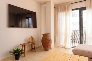 Ein Wohnzimmer mit Flachbildfernseher, einem Blumentopf, einem Holzschemel, einer großen Vase, einer Couch und einer Glastür zum Balkon.