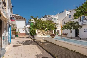 Une rue étroite bordée d’immeubles, avec une fontaine et des plantes en pot.