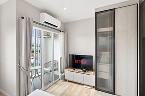 A modern living room with a sliding glass door leading to a balcony, a flat-screen TV on a wooden console, and a ceiling-mounted air conditioning unit. - Cozy 1-bedroom apartment in Title Serenity Naiyang (Thalang District)