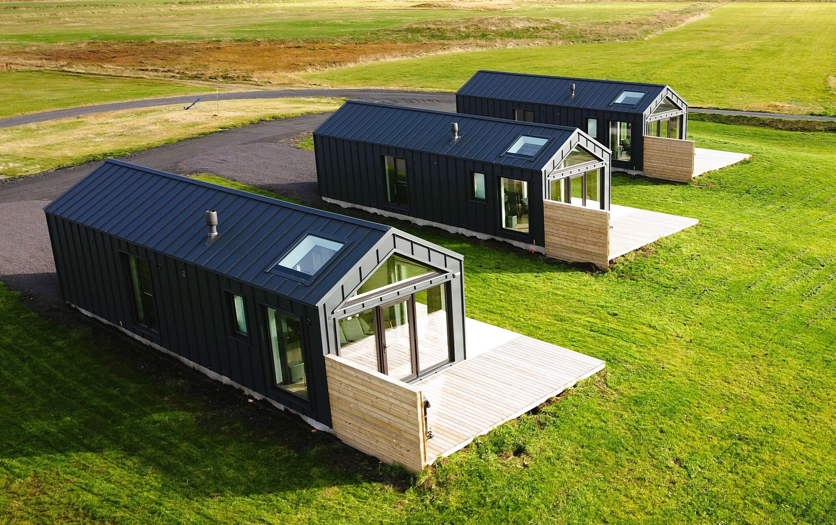 Tres cabañas modernas de color oscuro con grandes ventanales y terrazas de madera, ubicadas en un campo cubierto de hierba.