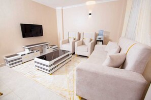 A modern living room with a flat-screen TV, a sofa, two armchairs, a coffee table, and a side table with a water dispenser. - Modern 1BR Apartment Near JKIA Balcony & Fast Wi-Fi (Nairobi)