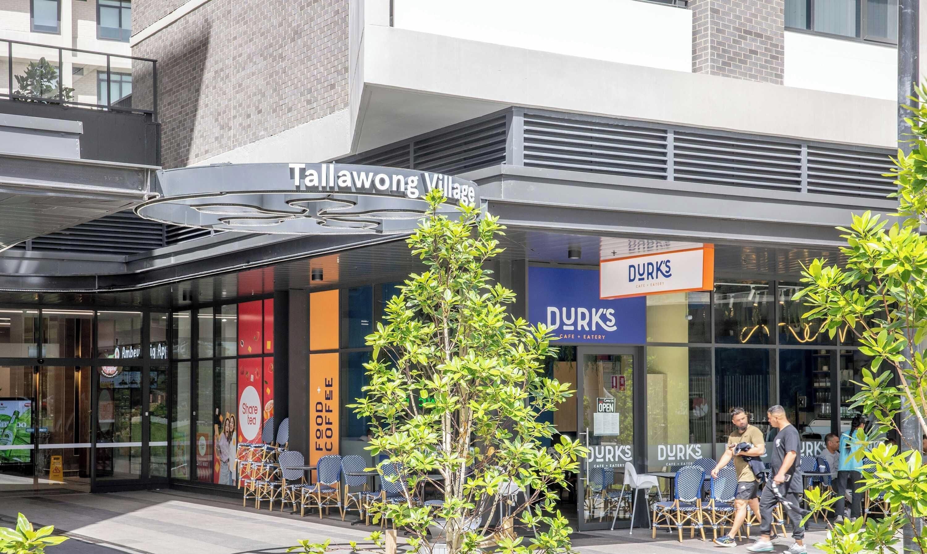 Un bâtiment moderne avec une façade en verre, un espace extérieur aménagé pour s’asseoir et des panneaux indiquant « Tallahong Village » et « DURK’S Cafe & Eatery ».