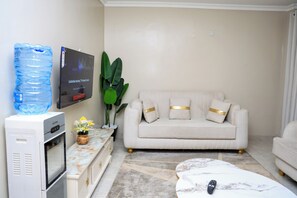 A living room with a flat-screen TV mounted on the wall, a sofa with decorative pillows, a coffee table, and a plant. - Cosy 1 Bedroom Apartment Near JKIA Airport Nairobi (Nairobi)
