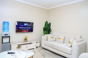 Ein Wohnzimmer mit Flachbildfernseher an der Wand, Wasserspender, Sofa mit goldenen Akzenten, kleiner Tisch mit Blumenvase und eine große Zimmerpflanze.
