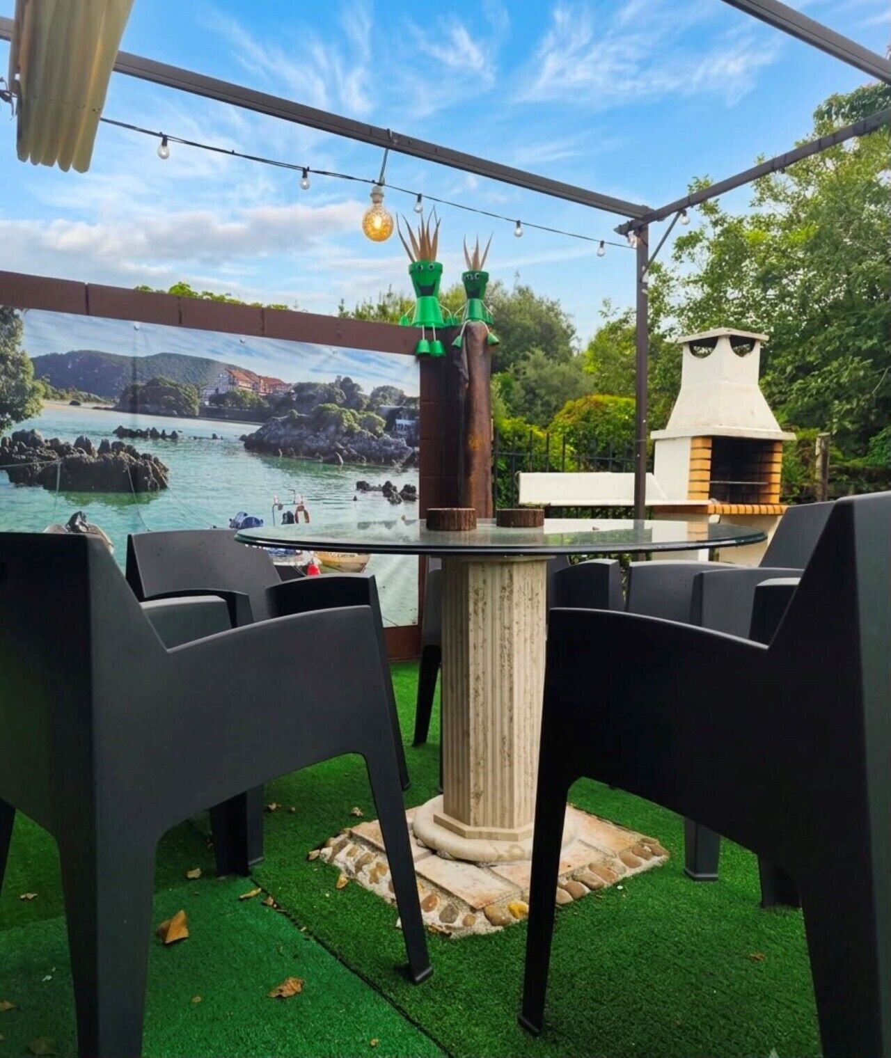 Un espacio al aire libre con mesas y vistas a un paisaje costero, una parrilla y luces colgantes.
