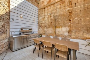 An outdoor kitchen with a grill, a dining table with chairs, and a stone wall backdrop. - 3BR Townhouse with Private Pool & BBQ in Birkirkara (Birkirkara)