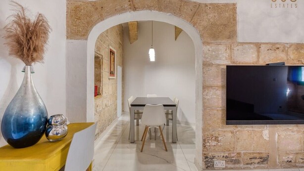A dining area with a stone archway, a table with chairs, a TV mounted on the wall, and a decorative vase on a yellow table. - 3BR Townhouse with Private Pool & BBQ in Birkirkara (Birkirkara)