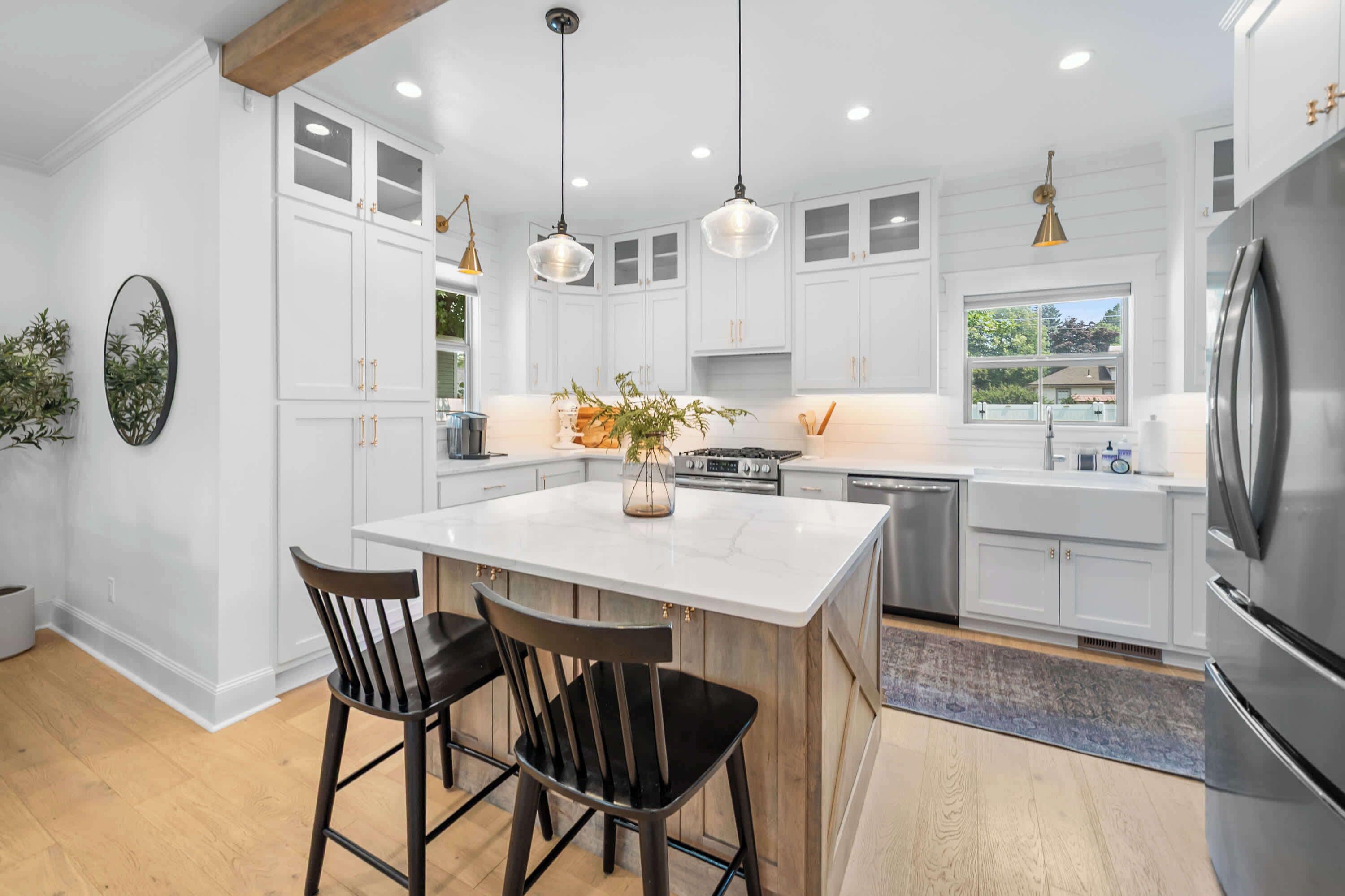 A modern kitchen with white cabinetry, a central island with two chairs, stainless steel appliances, pendant lighting, and a view to the outside.