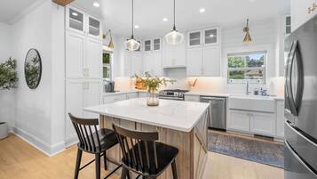 A modern kitchen with white cabinetry, a central island with two chairs, stainless steel appliances, pendant lighting, and a view to the outside.